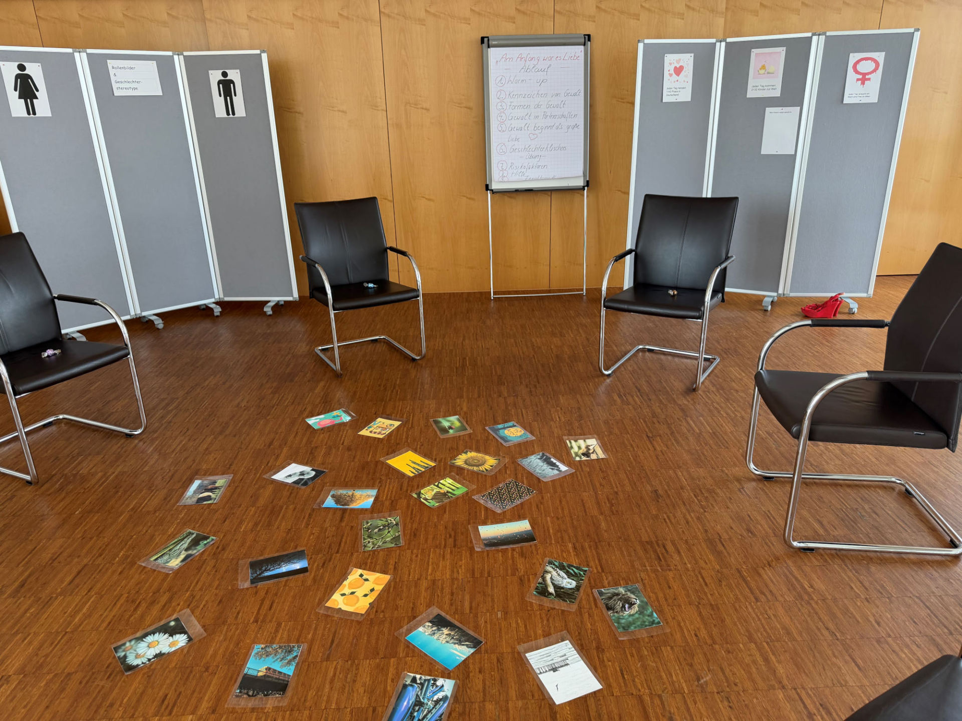 Es ist ein Stuhlkreis zu sehen, in dessen Mitte einige Fotos verstreut liegen. Es sind keine Personen auf dem Bild.