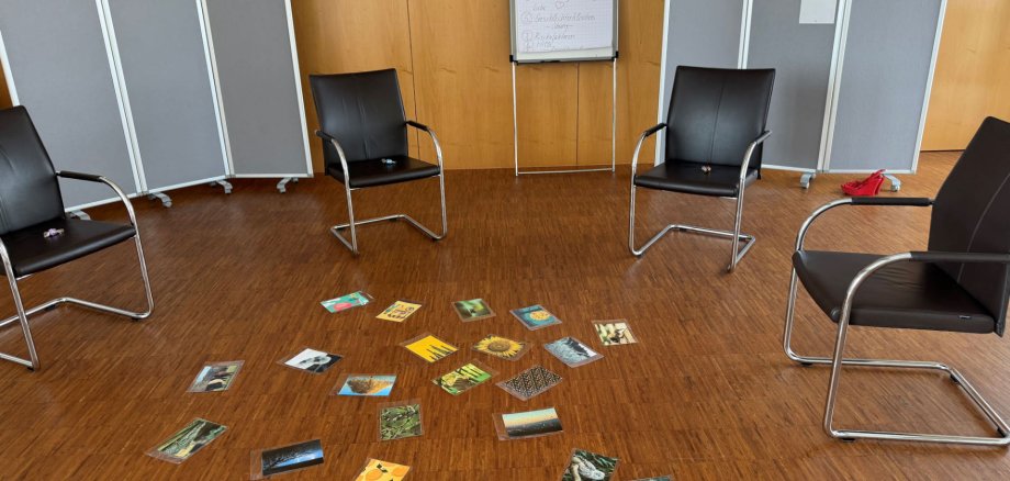 Es ist ein Stuhlkreis zu sehen, in dessen Mitte einige Fotos verstreut liegen. Es sind keine Personen auf dem Bild.