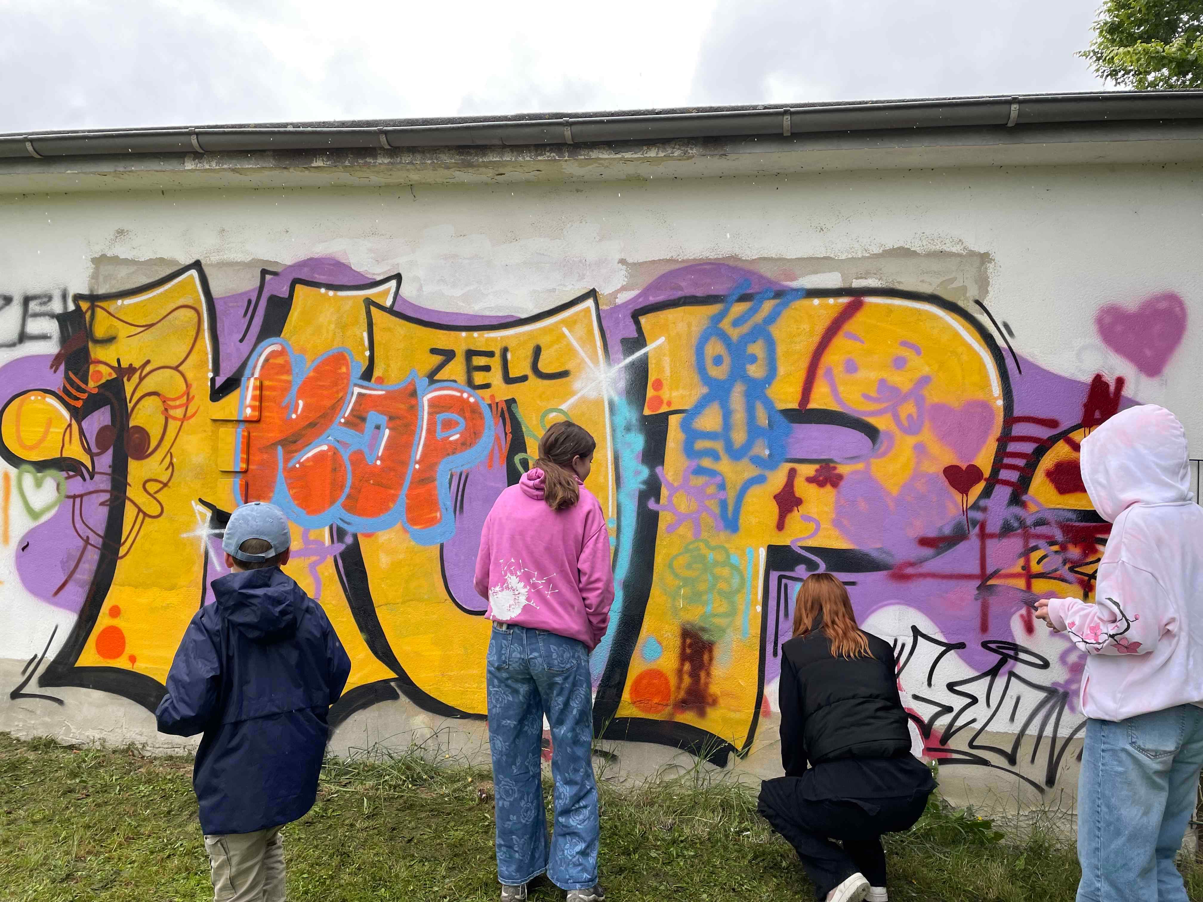 Vier Jugendliche stehen mit dem Rücken zur Kamera und sprühen bunte Graffiti an eine Wand. 