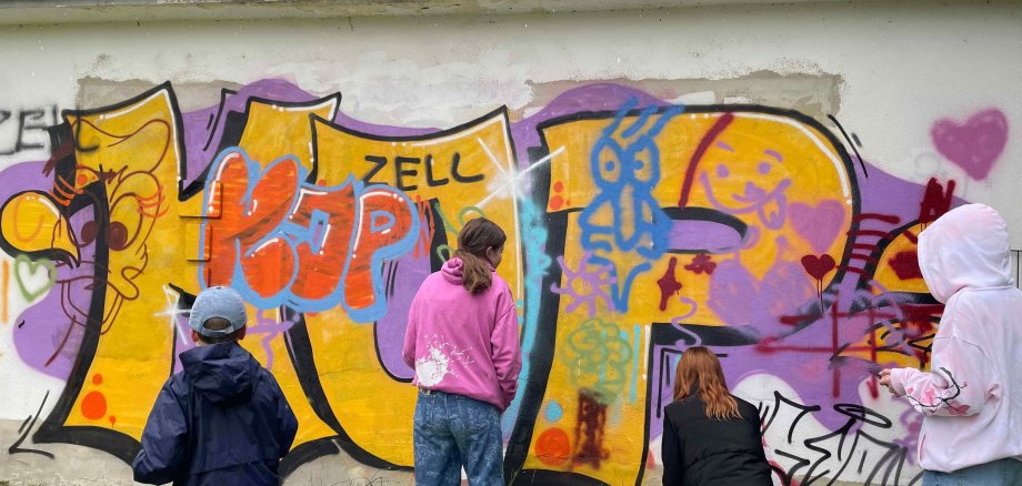 Vier Jugendliche stehen mit dem Rücken zur Kamera und sprühen bunte Graffiti an eine Wand. 