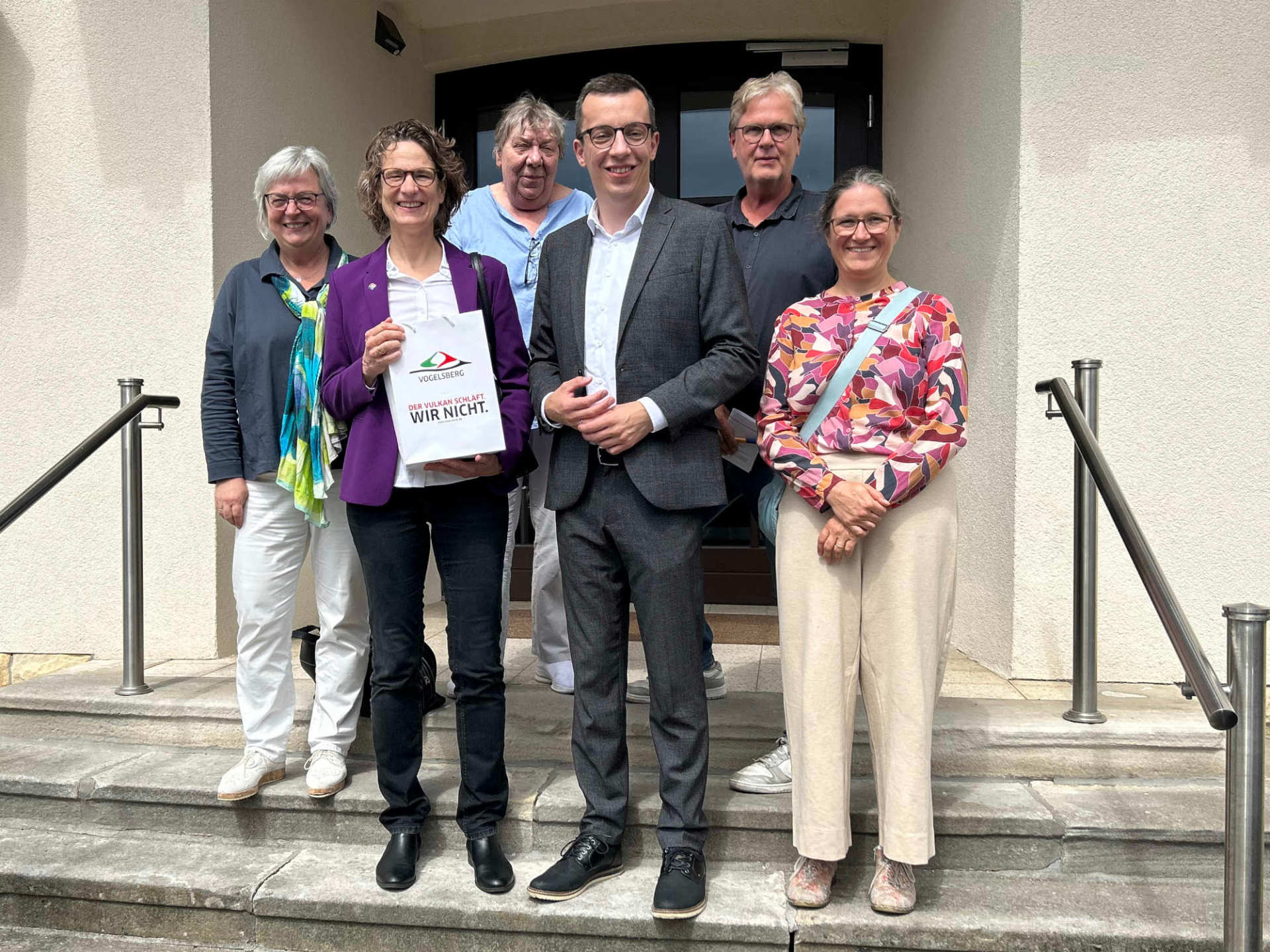 Sechs Personen haben sich auf einer Treppe für ein Gruppenfoto aufgestellt, und lächeln in die Kamera. Darunter sind auch Erster Kreisbeigeordneter Patrick Krug und Kirchenpräsidentin Prof. Dr. Christine Tietz.