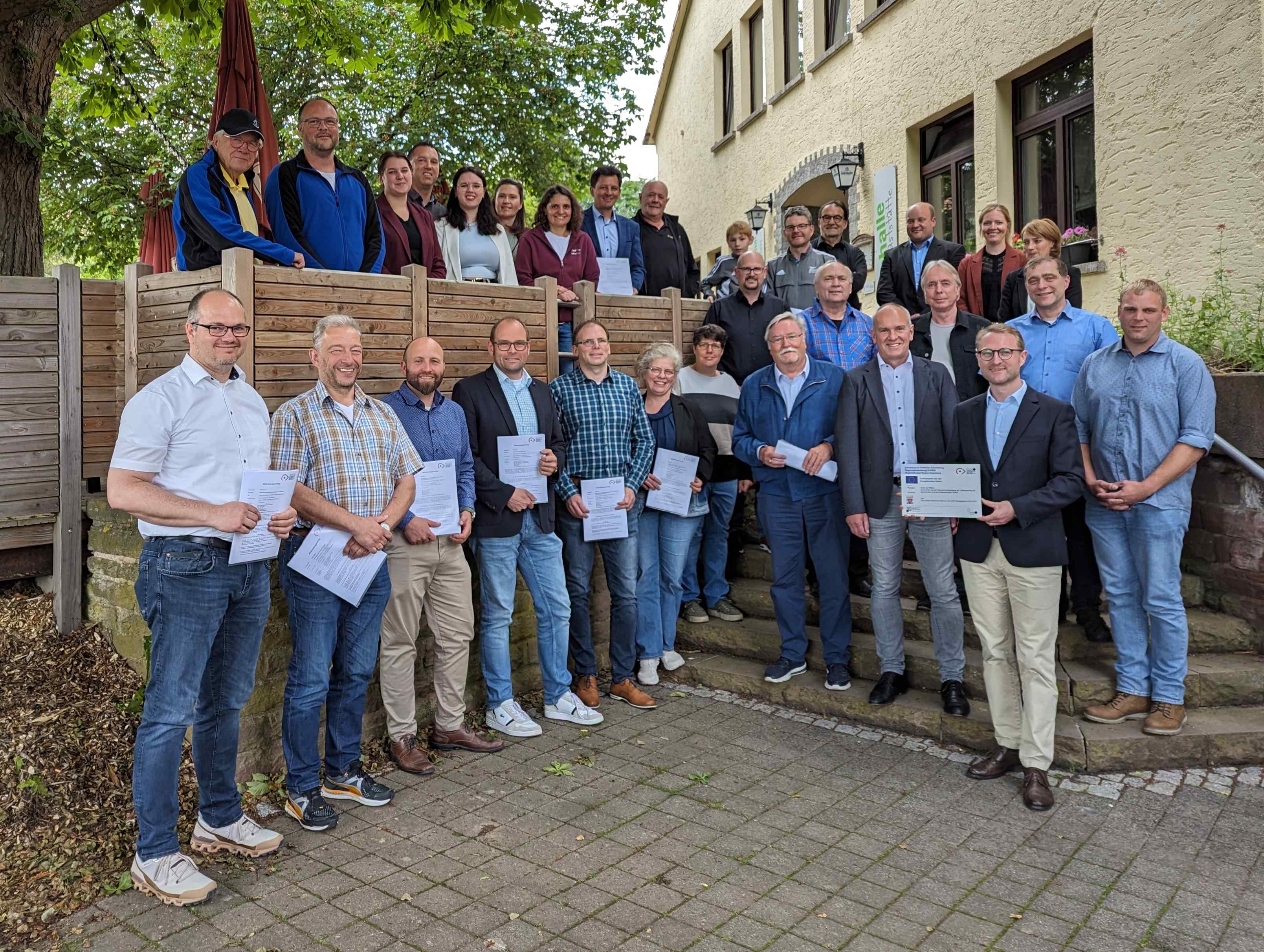 Dr. Jens Mischak (vorne, Zweiter von rechts), Vorstandsvorsitzender des Vereins Region Vogelsberg, und Edwin Schneider (Dritter von rechts), Vorsitzender des LEADER-Beirats, gemeinsam mit Vertreterinnen und Vertretern der Vereine und Kommunen sowie des LEADER-Regionalmanagements bei der Übergabe der Zuwendungsverträge an der Turnhalle Frischborn.