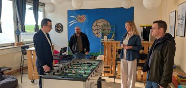 Vier Menschen stehen in einem Sozialraum einer Schule. In der Mitte steht ein Tisch-Kick-Gerät