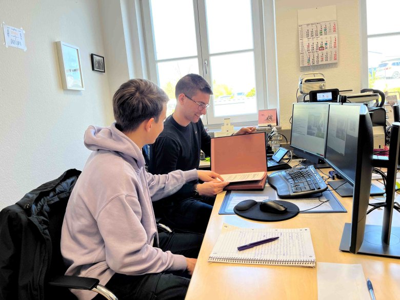 Paul Eidmann vom Personalservice (hinten) erläutert Leonas Pietsch seine Arbeit. Sie sitzen gemeinsam an einem Büroarbeitsplatz und schauen in eine Mappe