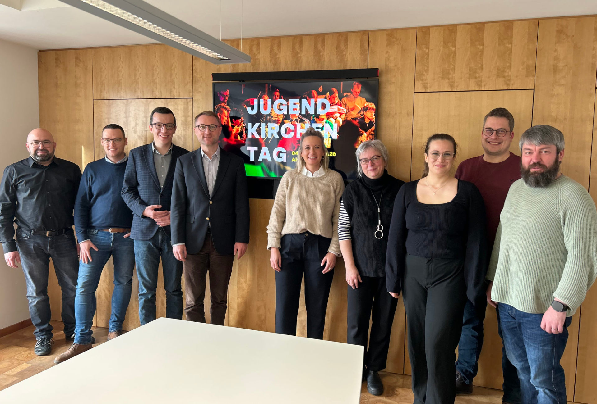 Mehrere Menschen haben sich in einem Konferenzraum für ein Gruppenbild aufgestellt. Sie lächeln in die Kamera und werben für den Jugendkirchentag 2026 in Alsfeld