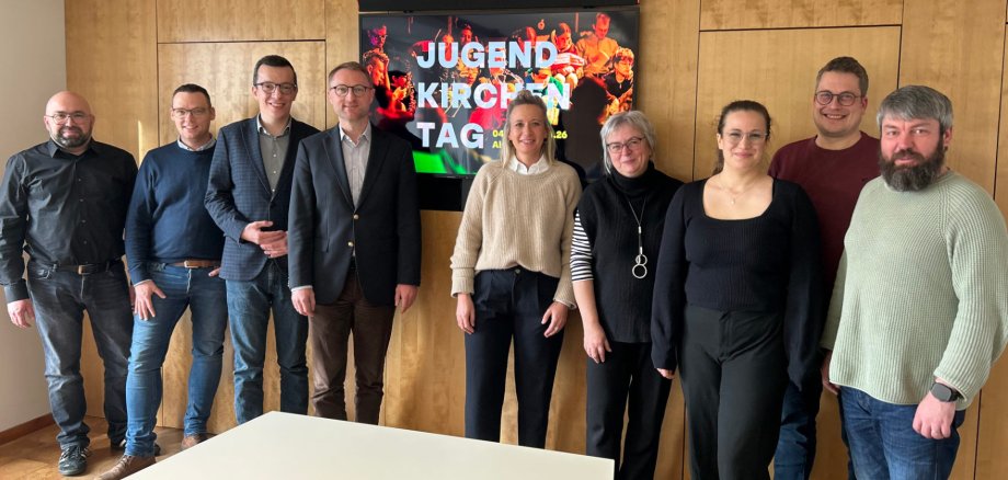 Mehrere Menschen haben sich in einem Konferenzraum für ein Gruppenbild aufgestellt. Sie lächeln in die Kamera und werben für den Jugendkirchentag 2026 in Alsfeld