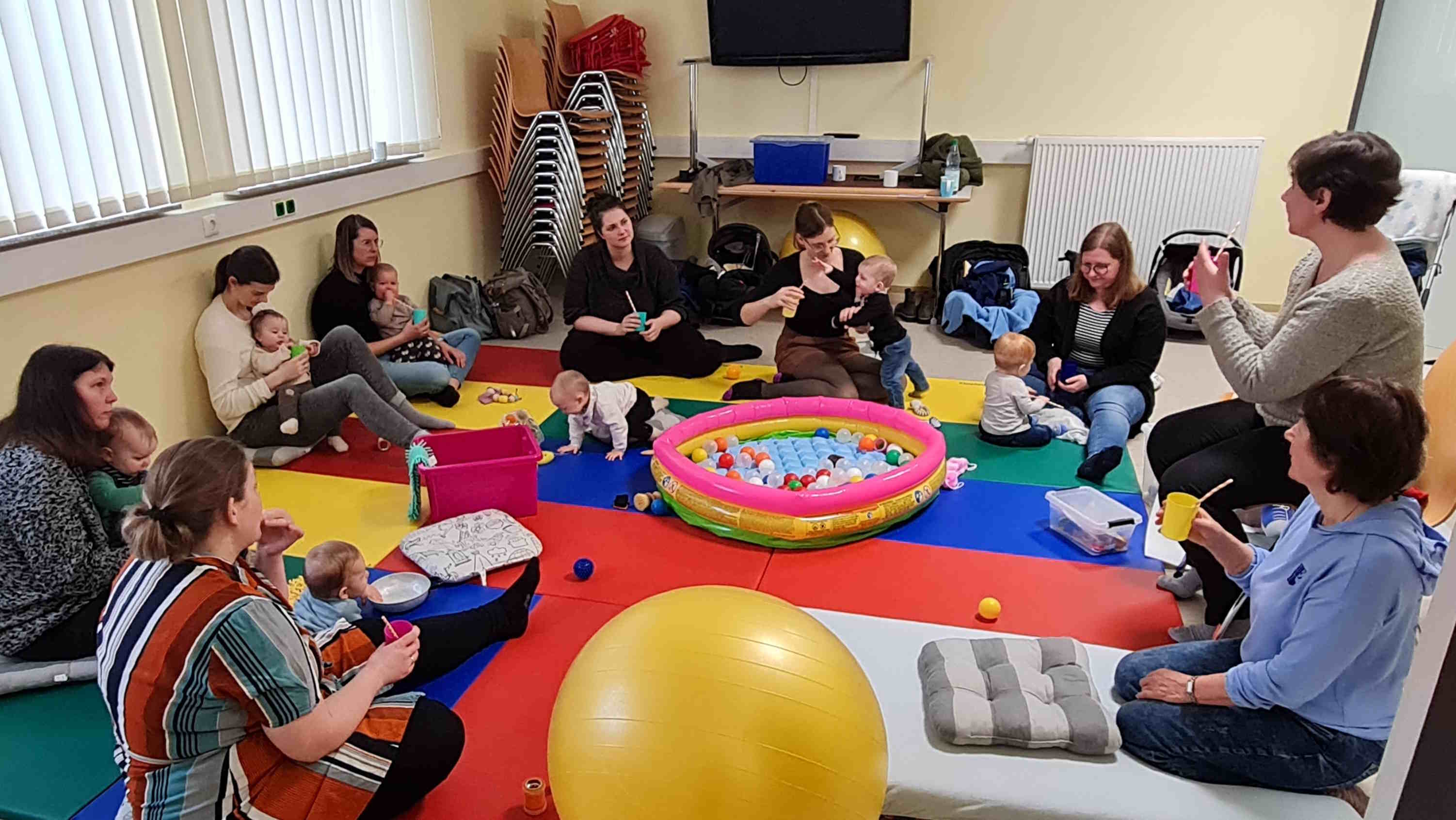 Mütter mit Kleinkindern sitzen auf dem Boden in einem Kreis. Sie hören einen anschaulichen Vortrag zum Thema Zahnpflege bei Kleinkindern und Babys