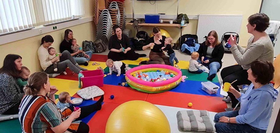 Mütter mit Kleinkindern sitzen auf dem Boden in einem Kreis. Sie hören einen anschaulichen Vortrag zum Thema Zahnpflege bei Kleinkindern und Babys
