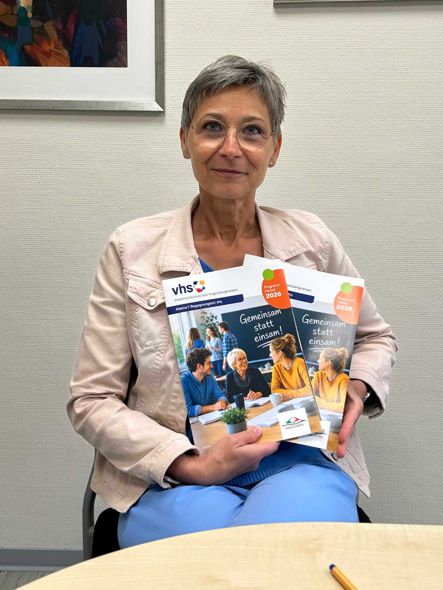 Kirsten Wegwerth sitzt an einem Tisch und präsentiert zwei Programmhefte. Sie lächelt in die Kamera