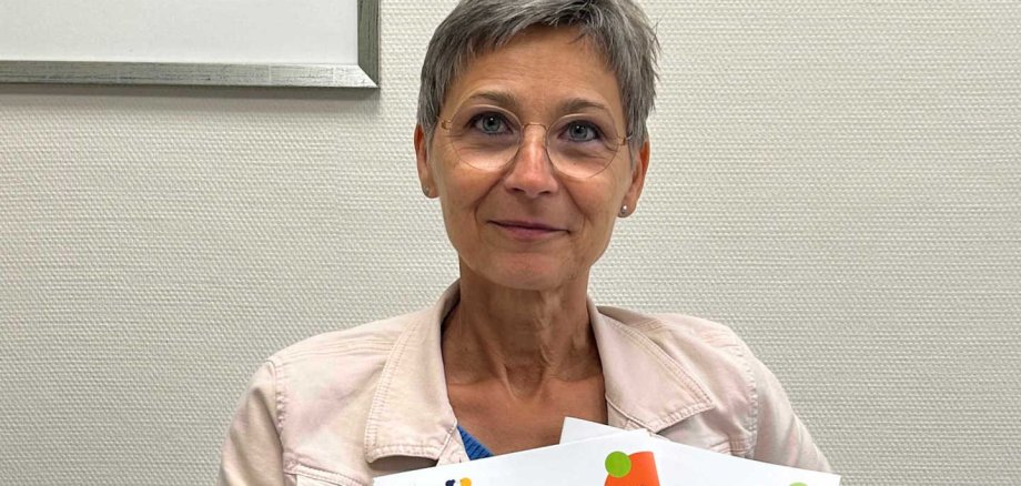 Kirsten Wegwerth sitzt an einem Tisch und präsentiert zwei Programmhefte. Sie lächelt in die Kamera