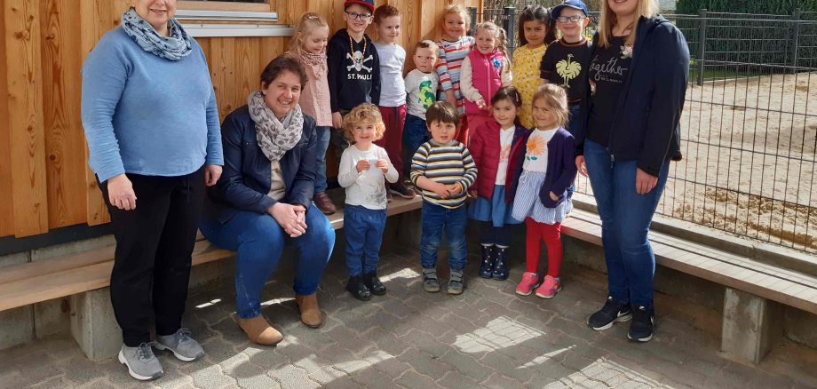 Gruppenbild in der Kita - viele Kita-Kinder stehen und sitzen vor einem kleinen Hölzhäuschen gemeinsam mit ihren Betreuerinnen.