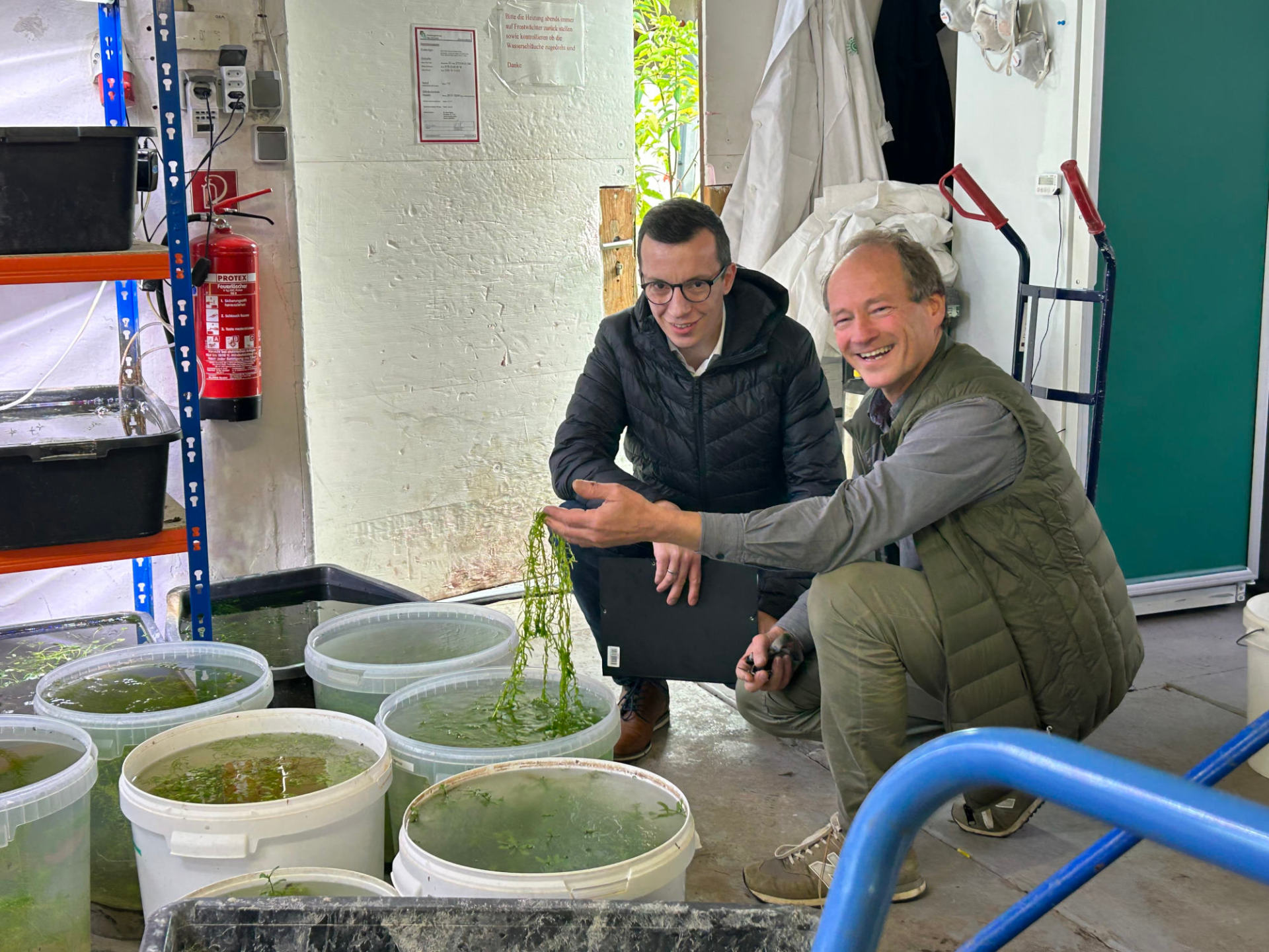Zwei Erwachsene hocken an Bottichen mit Experimenten und lächeln in die Kamera.