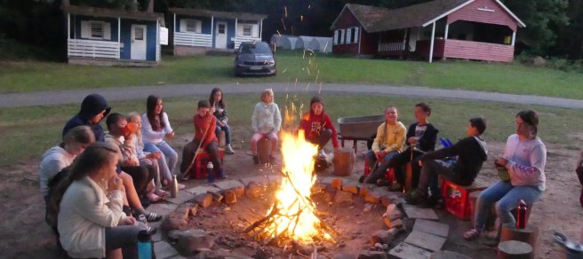 Etwa 15 Kinder sitzen im Kreis um ein Lagerfeuer. im Hintergrund sind kleine Häuschen zu sehen.