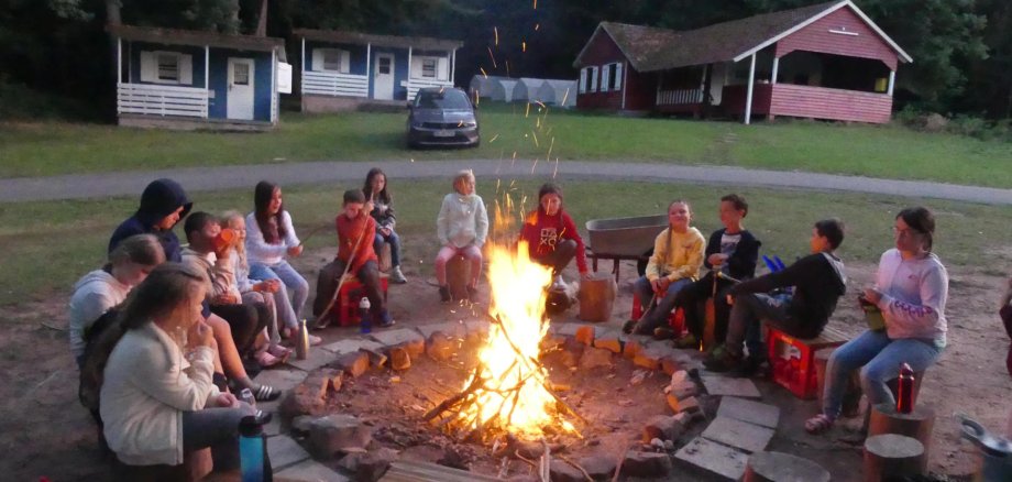 Etwa 15 Kinder sitzen im Kreis um ein Lagerfeuer. im Hintergrund sind kleine Häuschen zu sehen.