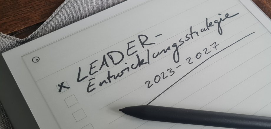Symbolfoto. ein Tablet liegt auf einem Holztisch. Das Tablet zeigt den handschriftlichen Text LEADER-Entwicklungsstrategie 2023-2027 an. 