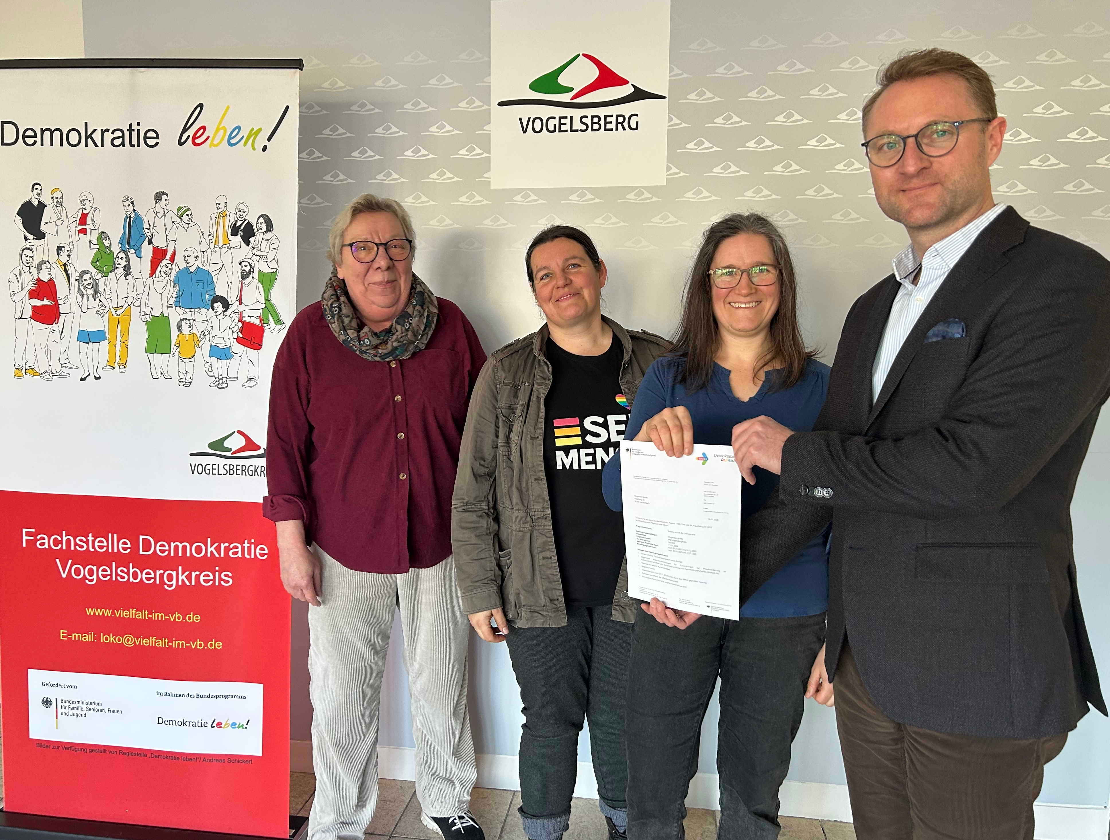 Vier Menschen stehen vor einer Fotowand und präsentieren einen Förderbescheid. Sie lächeln in die Kamera. 