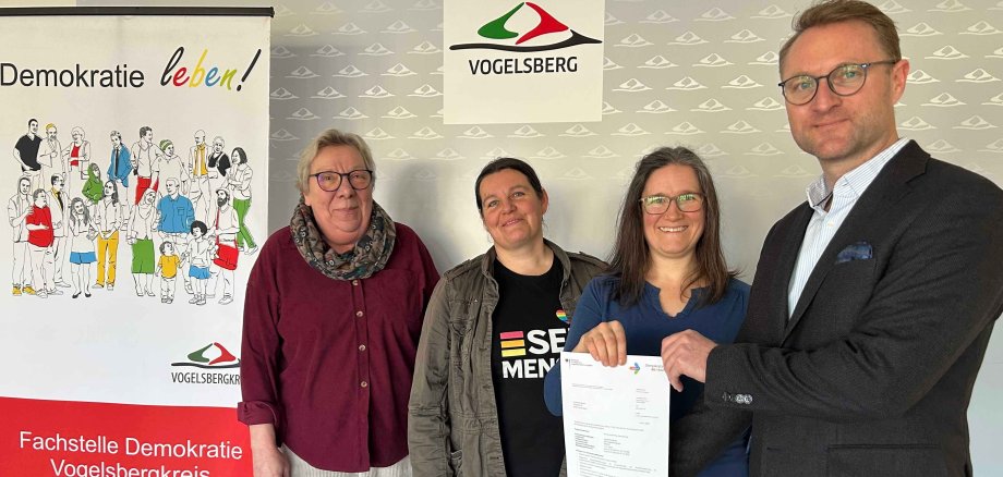 Vier Menschen stehen vor einer Fotowand und präsentieren einen Förderbescheid. Sie lächeln in die Kamera. 