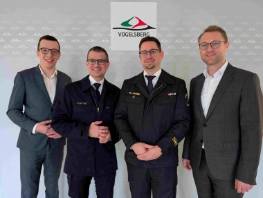 Vier Männer stehen vor einer grauen Wand. Zwei von ihnen tragen Feuerwehruniform