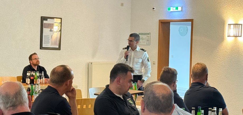 Ein Mann spricht mit Mikrofon in einem Bürgerhaus vor einer Gruppe von Menschen. 