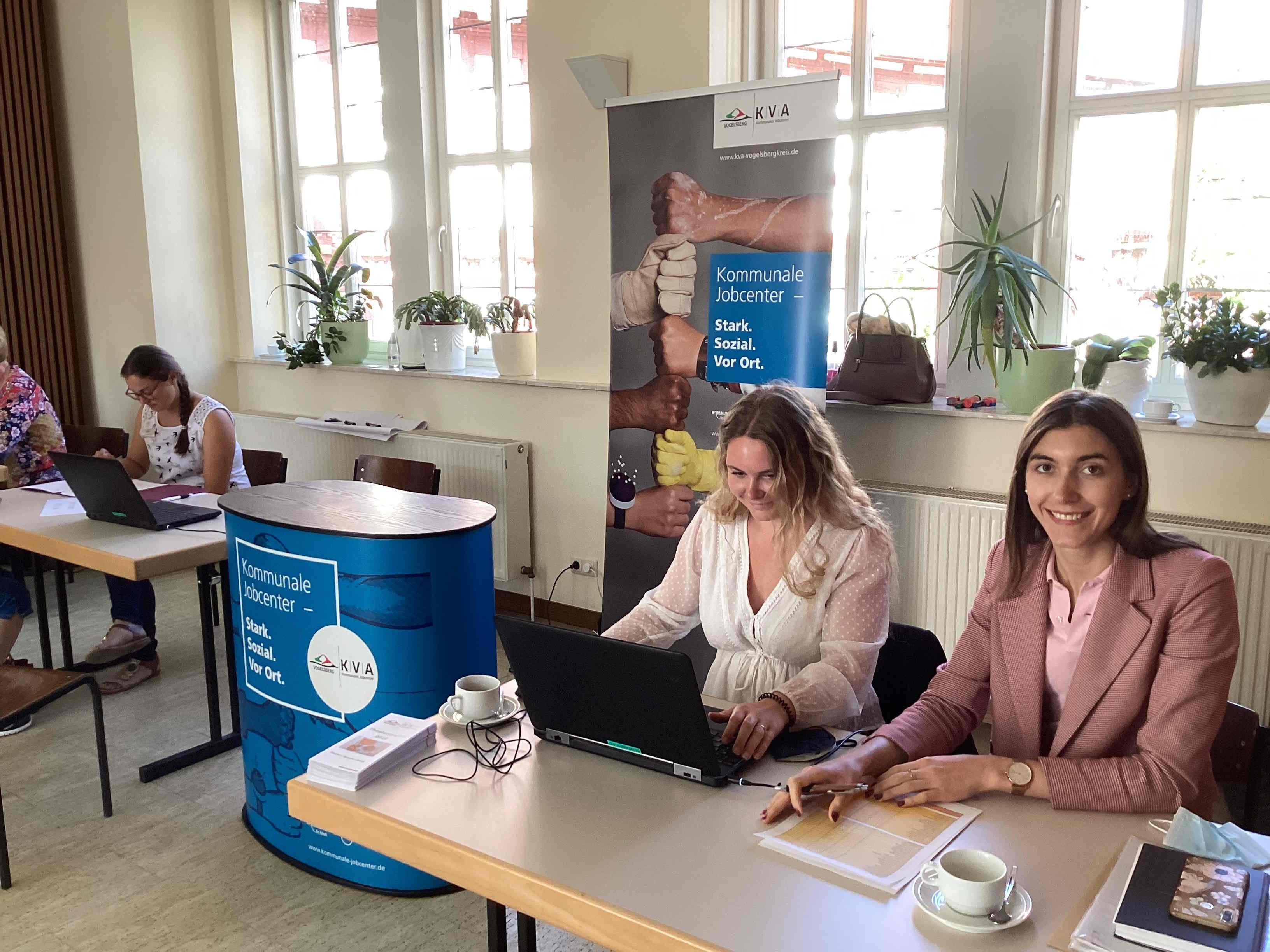 Zwei Mitarbeiterinnen der KVA Vogelsbergkreis – Kommunales Jobcenter beraten die Besucher bei der Minimesse in Schotten. Sie sitzen an einem Tisch und haben Unterlagen und einen Laptop vor sich.