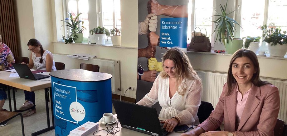 Zwei Mitarbeiterinnen der KVA Vogelsbergkreis – Kommunales Jobcenter beraten die Besucher bei der Minimesse in Schotten. Sie sitzen an einem Tisch und haben Unterlagen und einen Laptop vor sich.