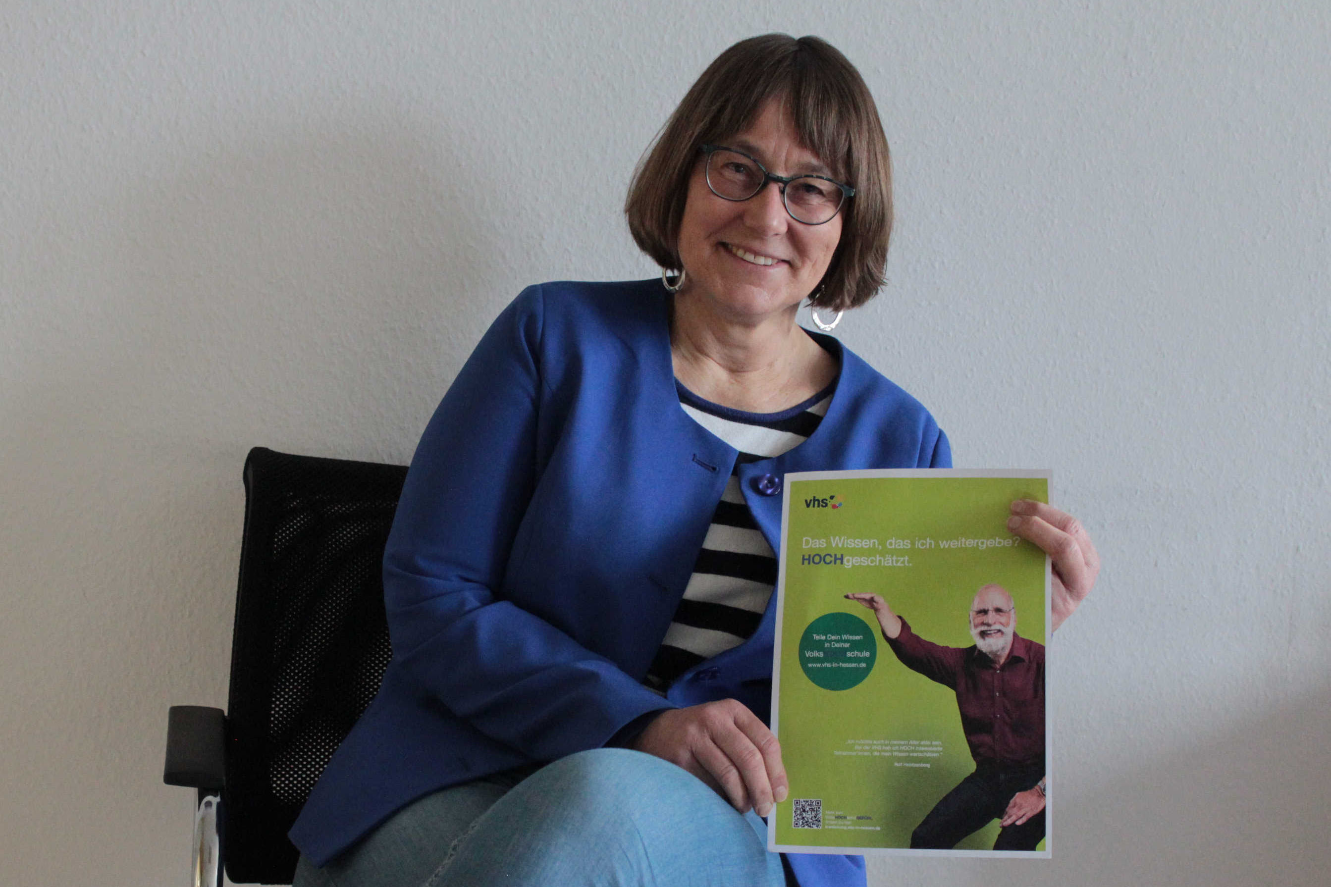 Monika Schenker sitzt auf einem Stuhl und hält ein Plakat in die Kamera. Sie wirbt für Kursleitungen an der vhs
