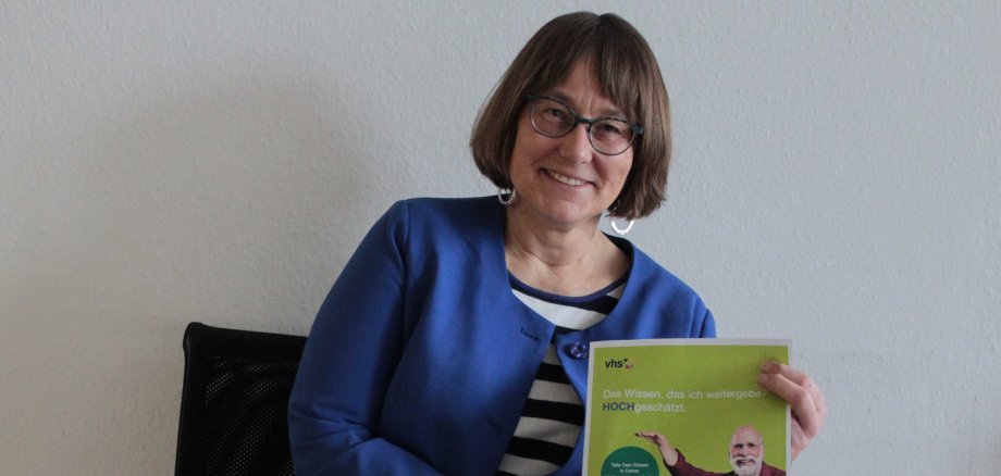 Monika Schenker sitzt auf einem Stuhl und hält ein Plakat in die Kamera. Sie wirbt für Kursleitungen an der vhs