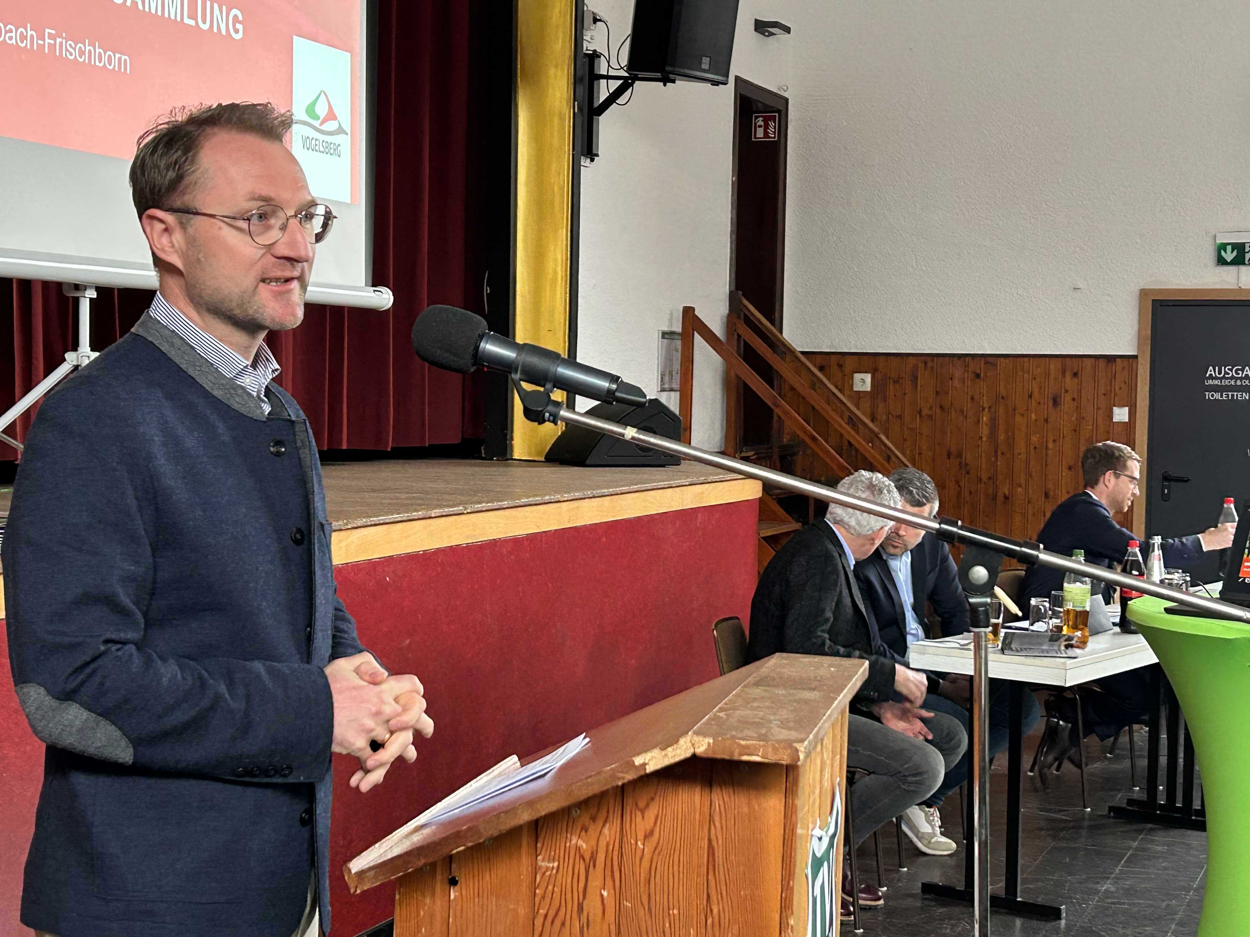 Landrat Dr. Mischak steht an einem Rednerpult mit Mikrofon. Im Hintergrund unterhalten sich zwei Männer an einem Tisch. 
