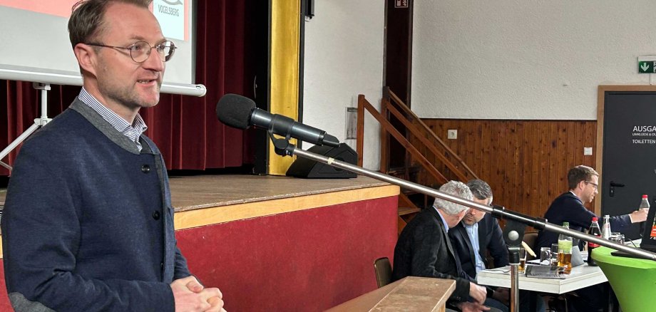 Landrat Dr. Mischak steht an einem Rednerpult mit Mikrofon. Im Hintergrund unterhalten sich zwei Männer an einem Tisch. 