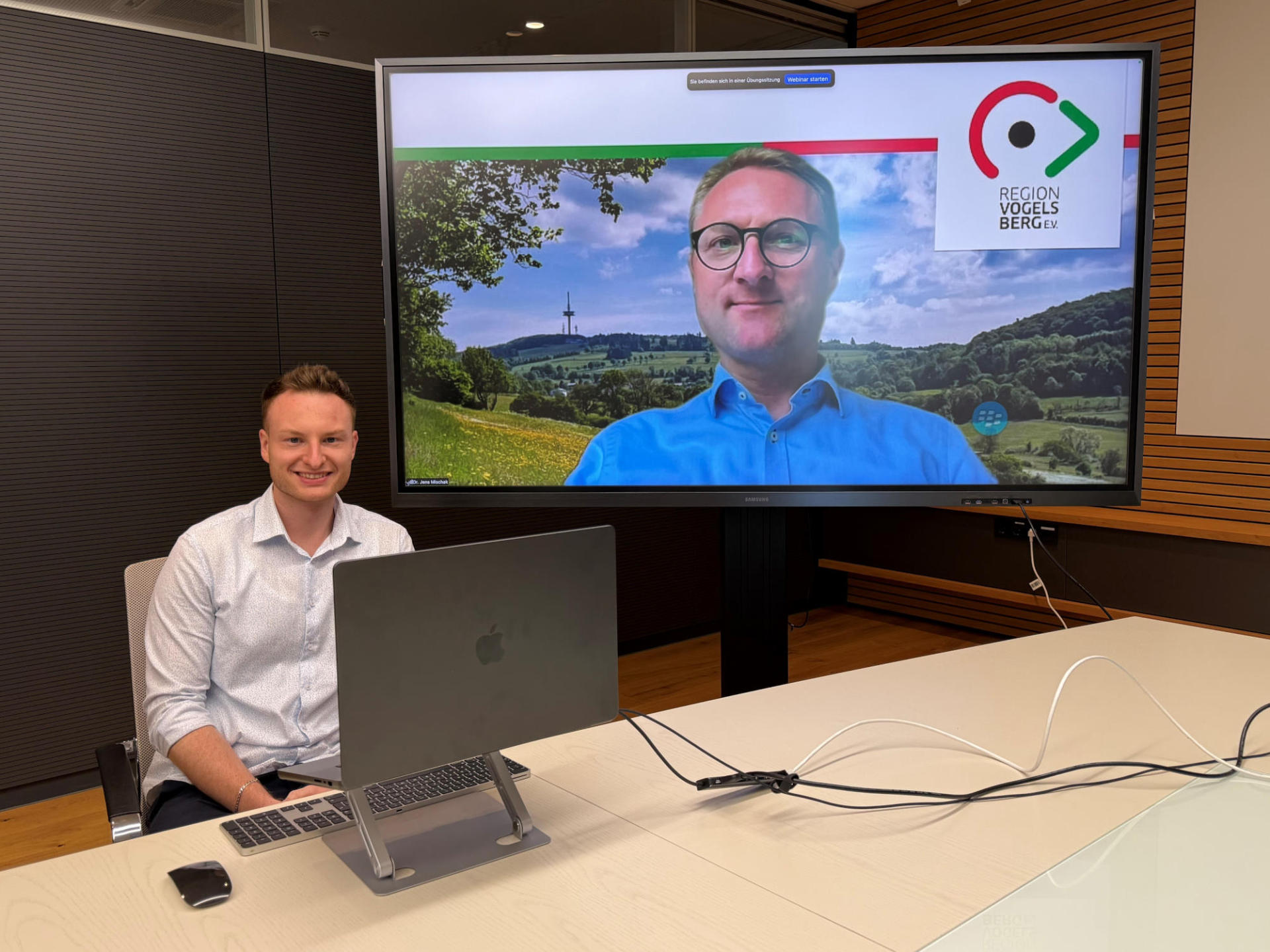 Ein Mann sitzt an einem Tisch und hat einen Laptop vor sich. Auf einem Bildschirm ist ein weiterer Mann zu sehen. 