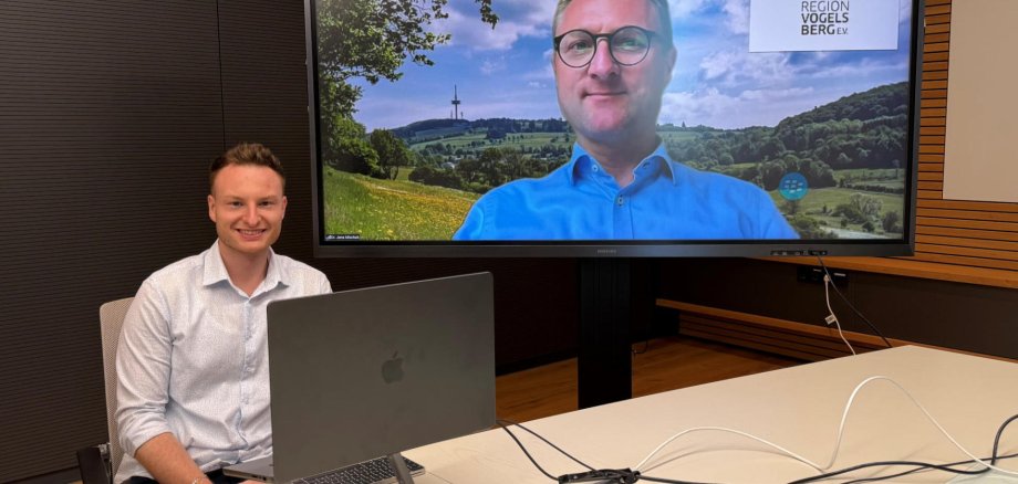 Ein Mann sitzt an einem Tisch und hat einen Laptop vor sich. Auf einem Bildschirm ist ein weiterer Mann zu sehen.