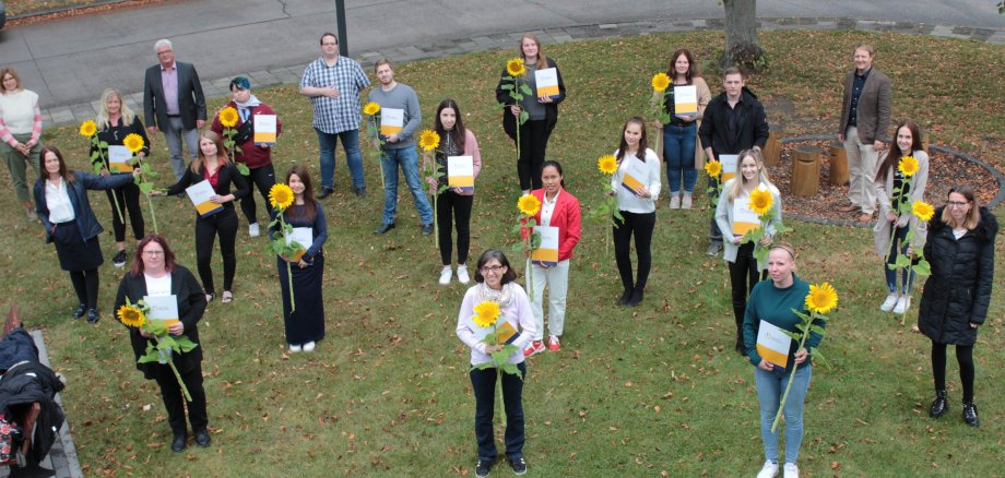 16 Altenpflegehelfer haben ihre einjährige Ausbildung an der Vogelsberger Pflegeakademie erfolgreich abgeschlossen. Sie stehen mit Sonnenblumen und ihren ZEugnissen vor der Pflegeakademie