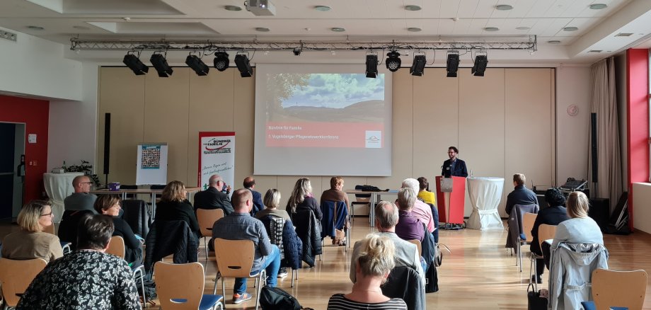 Simon Päbler, Sprecher des Familienbündnisses Vogelsberg, moderiert die 1. Vogelsberger Pflegenetzwerkkonferenz und führt durch den Nachmittag in der Aula der Alexander-von-Humboldt-Schule in Lauterbach. Er steht an einem Rednerpult und hält vor etwa 30 Menschen einen Vortrag. 