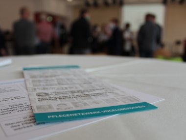 Ein Bild von einem Flyer in der Nahaufnahme. Darauf ist Pflegenetzwerk Vogelsbergkreis zu lesen. im Hintergrund sind verschwommen Menschen zu sehen.