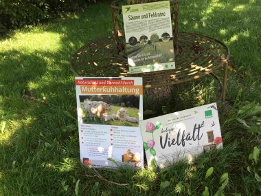 Drei Schilder zu Nachhaltigkeitsprojekten sind unter einem Baum aufgestellt. Es geht um Insektenschutz, vielfältige Pflanzenarten und biologische Landwirtschaft