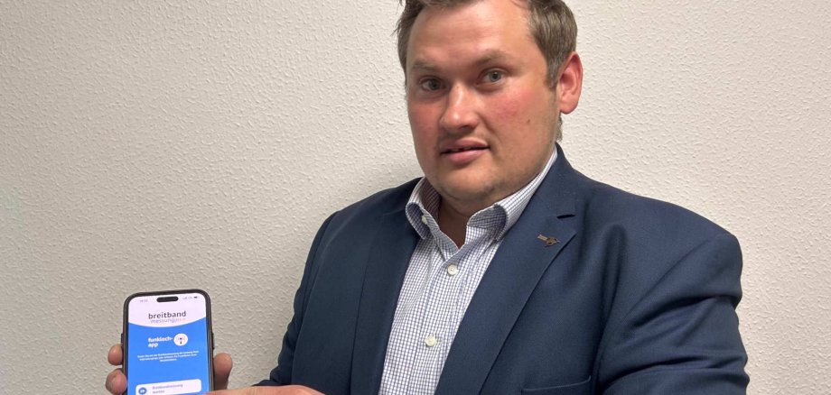 Ein Mann steht vor einer weiß gestrichenen Wand und deutet auf sein Handy. Er macht auf einen Aktionstag aufmerksam.