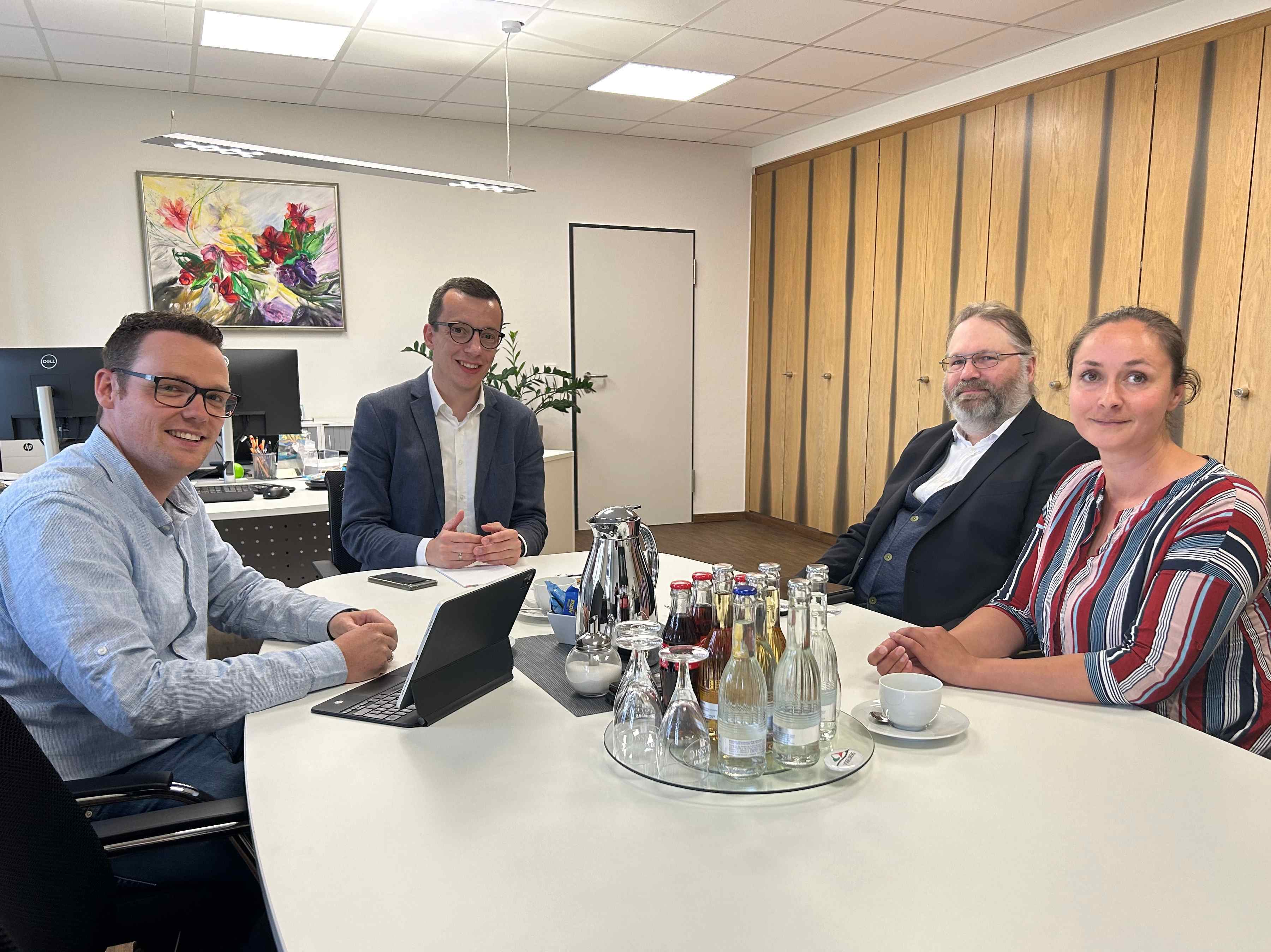 Vier Menschen sitzen an einem Tisch und schauen in die Kamera. Sie sitzen in einem Büro.