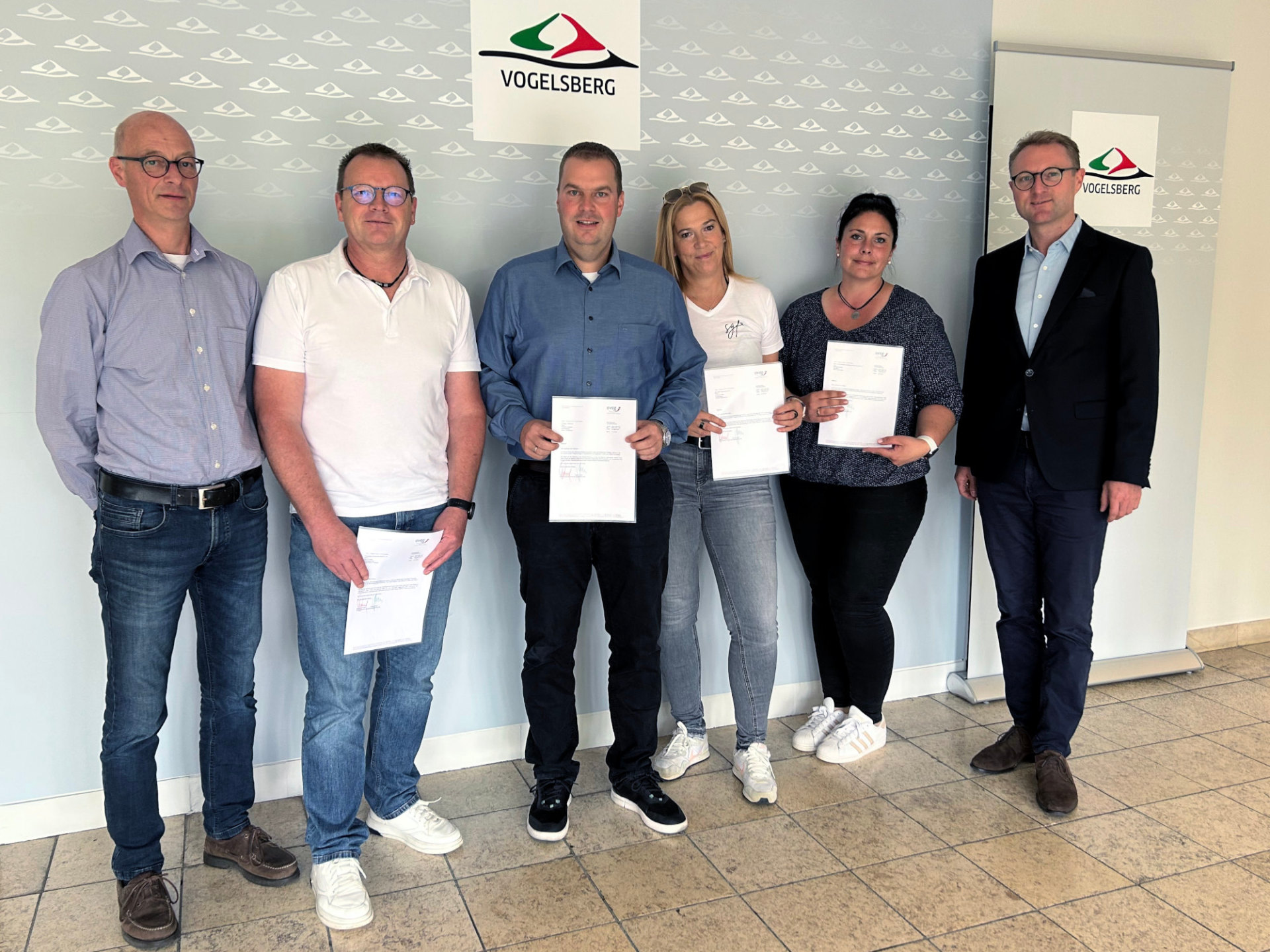 Sechs Personen haben sich vor einer Fotowand aufgestellt. Sie lächeln in die Kamera und halten Spendenbescheide in die Kamera.