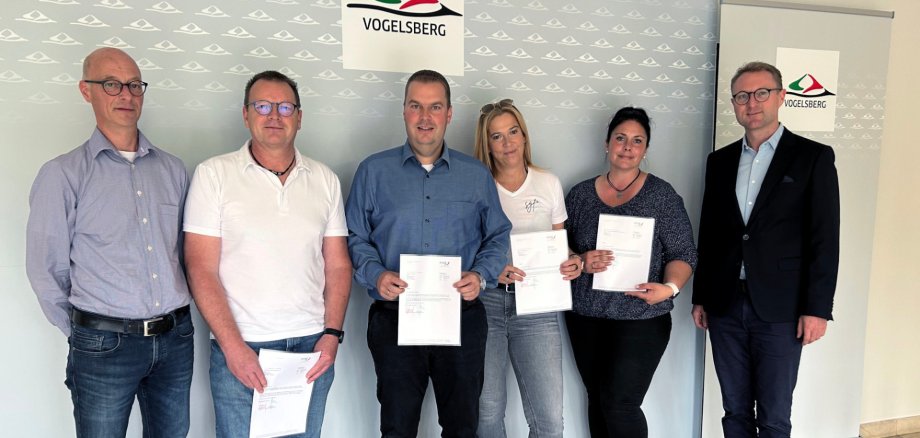 Sechs Personen haben sich vor einer Fotowand aufgestellt. Sie lächeln in die Kamera und halten Spendenbescheide in die Kamera.