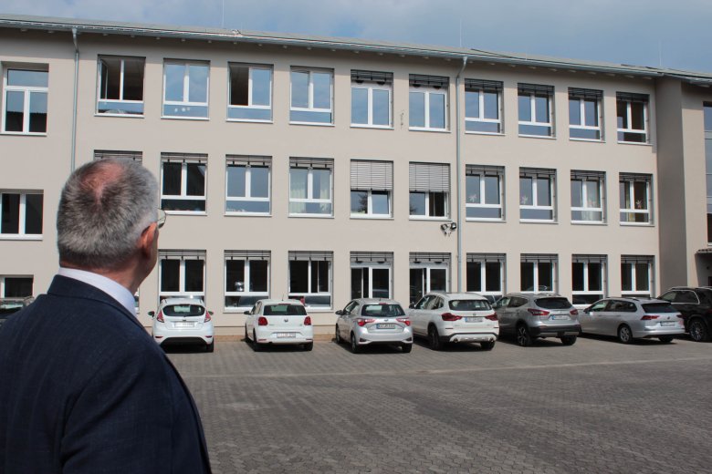 Landrat Manfred Görig steht mit dem Rücken zur Kamera vor einer Hausfassade eines Schulgebäudes. Diese ist frisch renoviert, vor dem Schulgebäude stehen Autos. 