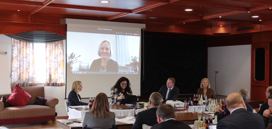 Vor einer Videoleinwand sitzt ein Podium. Darauf: Anita Schneider (von rechts), Landrätin des Landkreises Gießen, stellvertretende Aufsichtsratsvorsitzende der VVT GmbH, Dr. Jens Mischak, Erster Kreisbeigeordneter des Vogelsbergkreises und Aufsichtsratsvorsitzender der VVT GmbH, Petra Schwing-Döring, Geschäftsführerin VVT GmbH, nehmen zum Auftakt der Sitzung gemeinsam mit Ilka Schacht, Mitarbeiterin bei der VVT GmbH, die guten Neuigkeiten von Yvonne Haider, Geschäftsführerin Tourismus-Management Hessen (TMH), die per Videokonferenz zugeschaltet war, entgegen.
