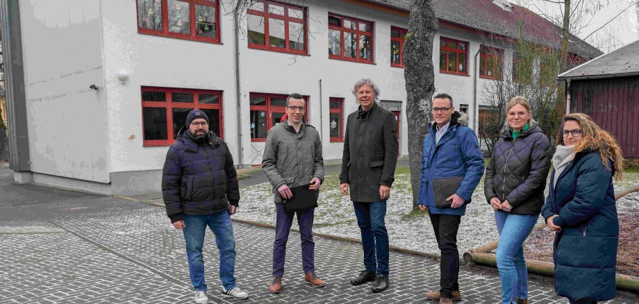 Sechs Menschen stehen vor einem Schulgebäude und lächeln in die Kamera. 