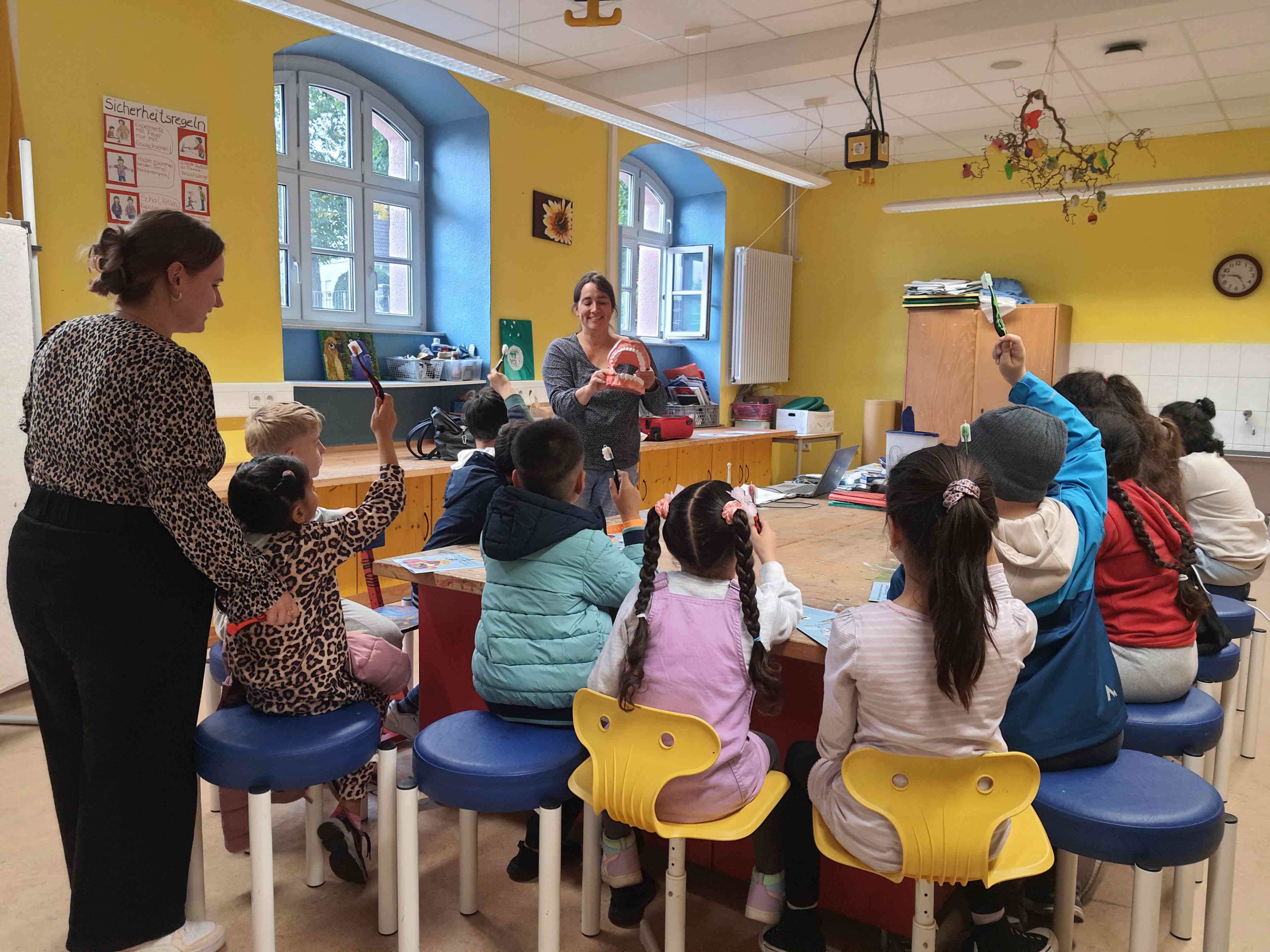 Eine Schulklasse lernt im Unterricht die richtige Zahnputztechnik gemeinsam mit einer Zahnärztin. Sie zeigt die Technik an einem großen künstlichen Gebiss.