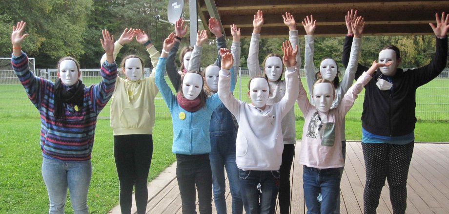 Auf dem Foto sind Kinder mit weißen  Masken zu sehen. Sie stehen auf einer Veranda und reißen die Arme in die Luft. Im Hintergrund ist ein Sportplatz und Wald zu sehen