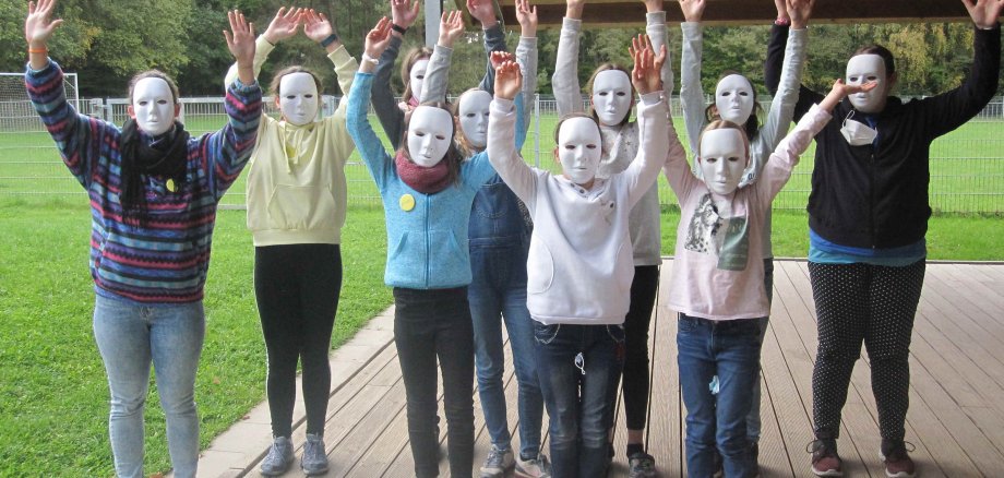 Auf dem Foto sind Kinder mit weißen  Masken zu sehen. Sie stehen auf einer Veranda und reißen die Arme in die Luft. Im Hintergrund ist ein Sportplatz und Wald zu sehen