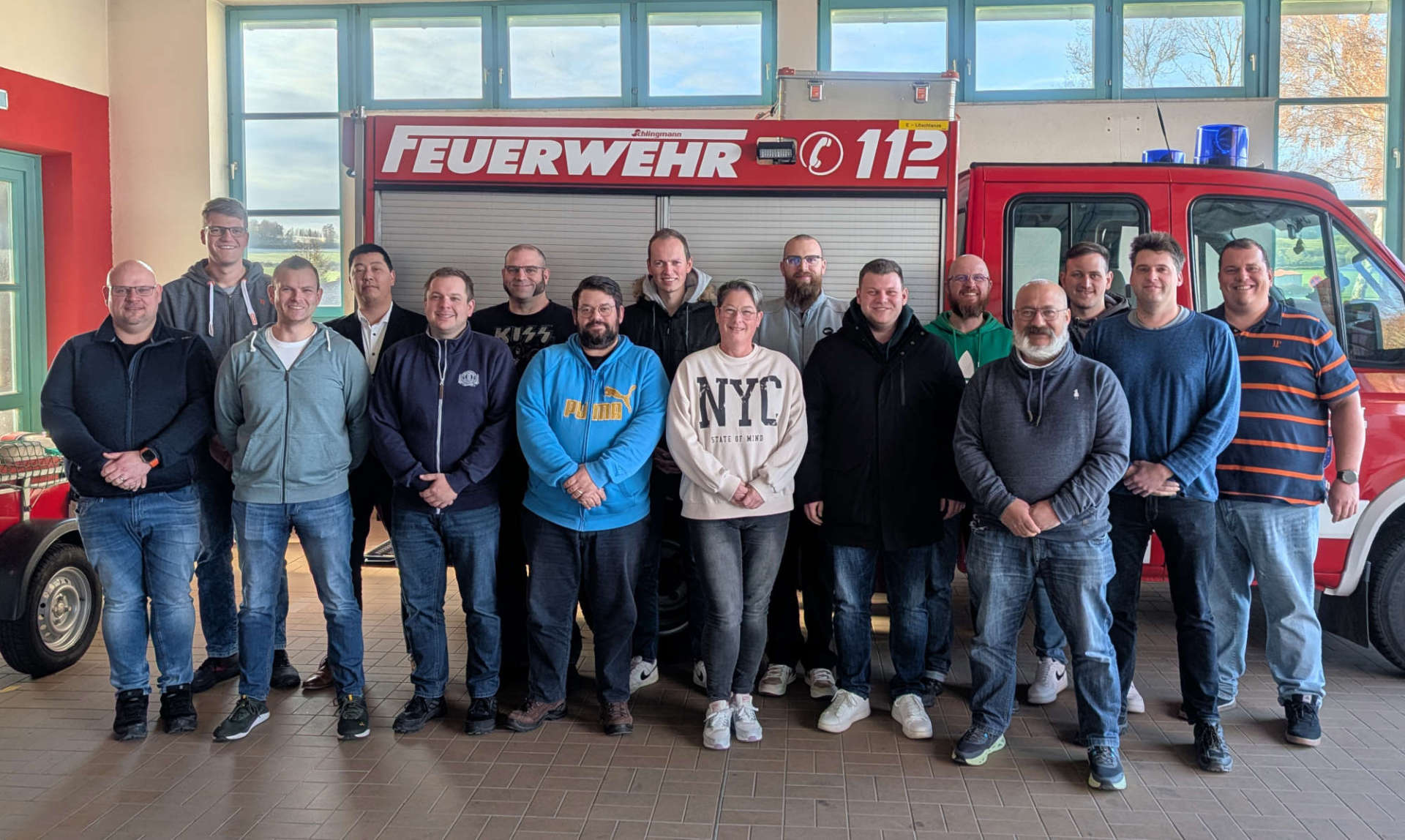 16 Personen haben sich vor einem Feuerwehrauto zum Gruppenbild aufgestellt und lächeln in die Kamera