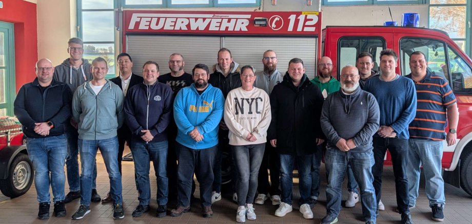 16 Personen haben sich vor einem Feuerwehrauto zum Gruppenbild aufgestellt und lächeln in die Kamera