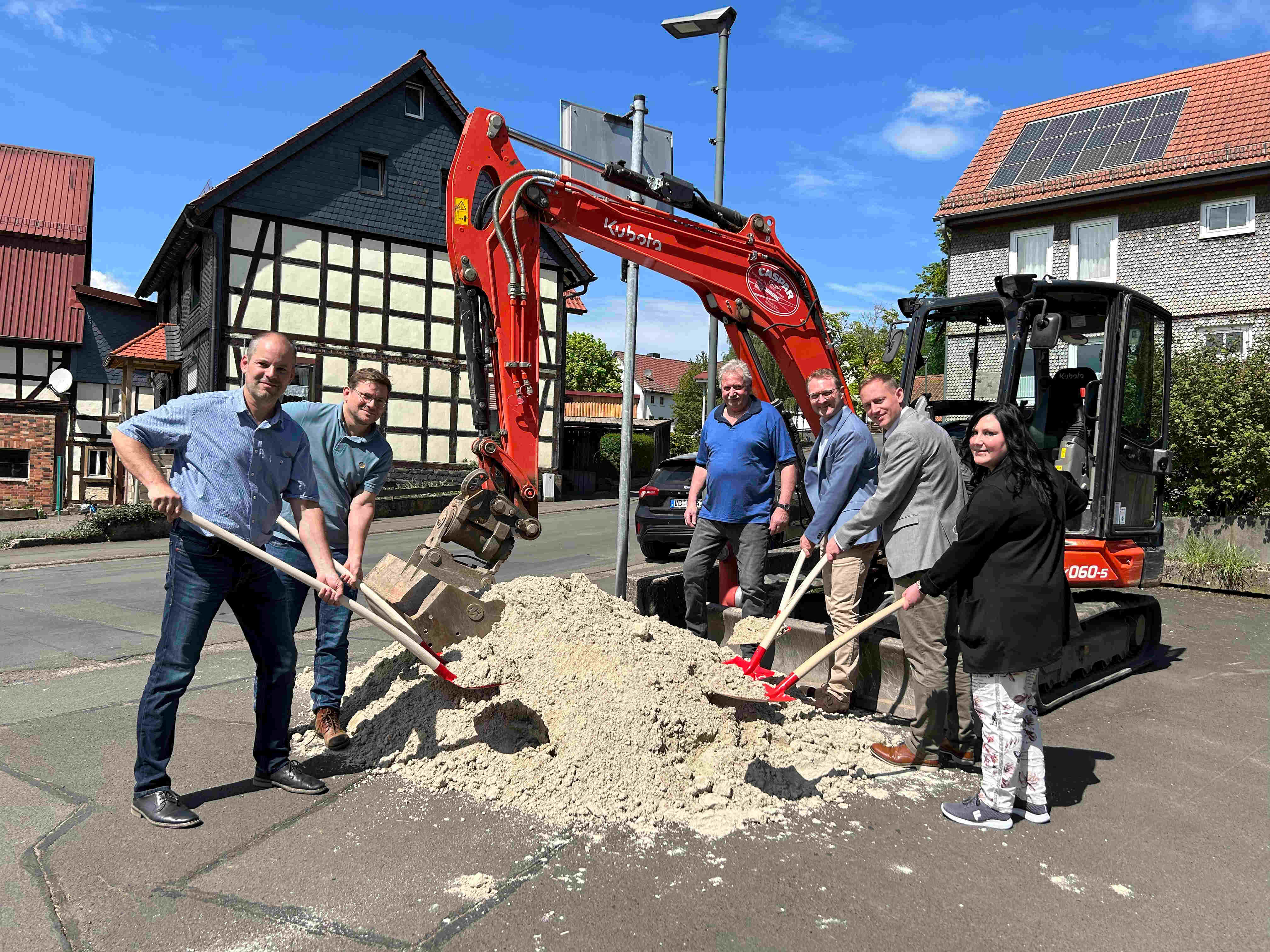 Sechs Menschen stehen an einem Sandhaufen vor einem kleinen Bagger. Sie führen symbolisch den Spatenstich für den Ausbau einer KReisstraße durch.