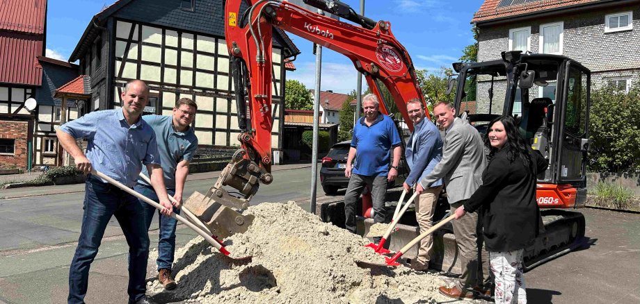 Sechs Menschen stehen an einem Sandhaufen vor einem kleinen Bagger. Sie führen symbolisch den Spatenstich für den Ausbau einer KReisstraße durch.