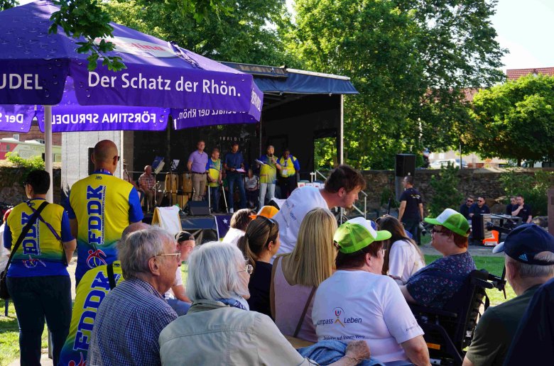 Einige Menschen sitzen an Bierzeltgarnituren in einem großen Garten. Sie schauen auf eine Bühne